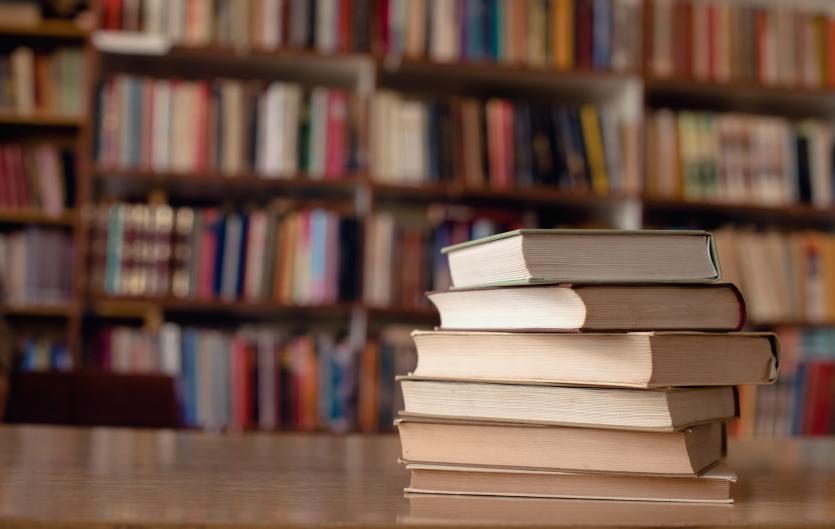 A stack of books on a table in front of bookshelves.
