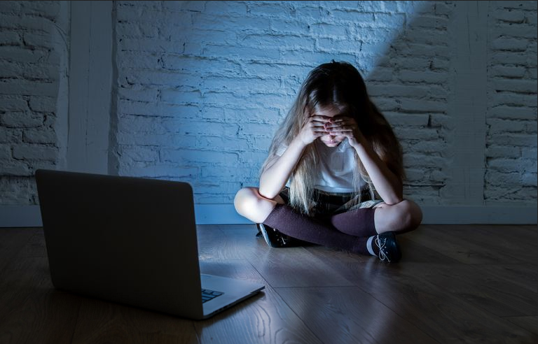 A young girl sits in a darkened room on the floor with her heads in her hands in front of an open laptop.