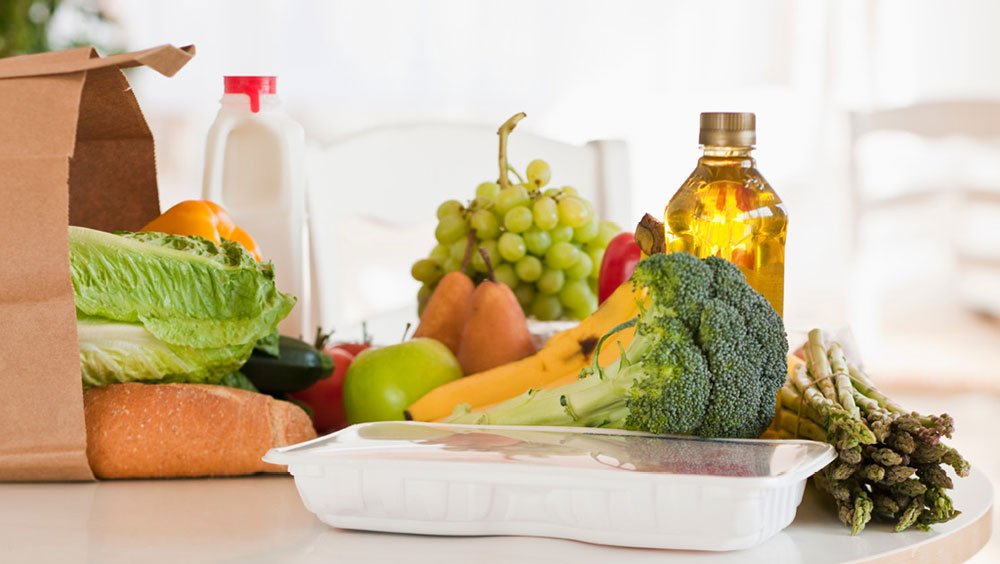Groceries on a counter, including a package of meat, jug of milk, container of oil, and fresh vegetables coming out of a paper bag.